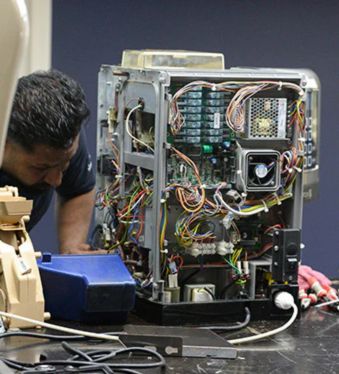 a technician leaning over, focused on repairing the intricate internal components of a large, complex coffee machine. The machine's casing is open, revealing a dense network of wires, circuit boards, fans, and other electronic and mechanical parts. The overall scene suggests a specialized repair workshop. The filename, like the previous one, indicates this is likely related to coffee machine repair services in Dubai.