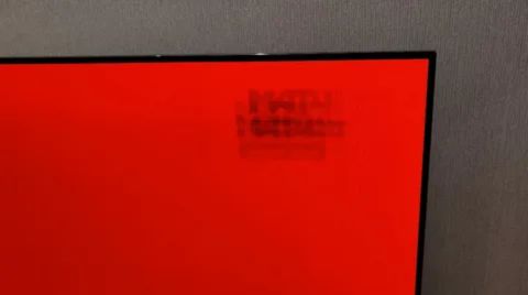 close-up shot of a screen (likely OLED) displaying a solid red color, specifically focusing on the top right corner.
The image clearly illustrates an instance of screen burn-in (image retention). A faint but permanently etched dark pattern, appearing to be text or a logo, is visible against the red background.